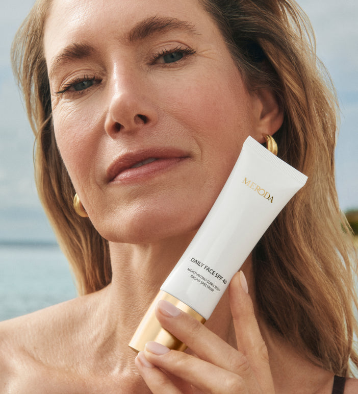 Model with radiant skin holding the Meroda Cosmetics Daily Face SPF 40 tube in a sunny beach setting.