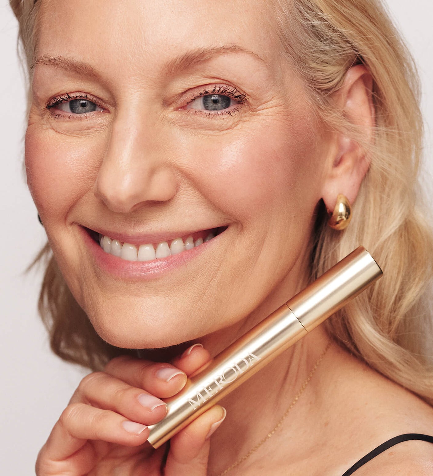 Smiling mature woman holding Meroda Cosmetics Gilded Lashes Mascara to showcase lengthened and volumized lashes