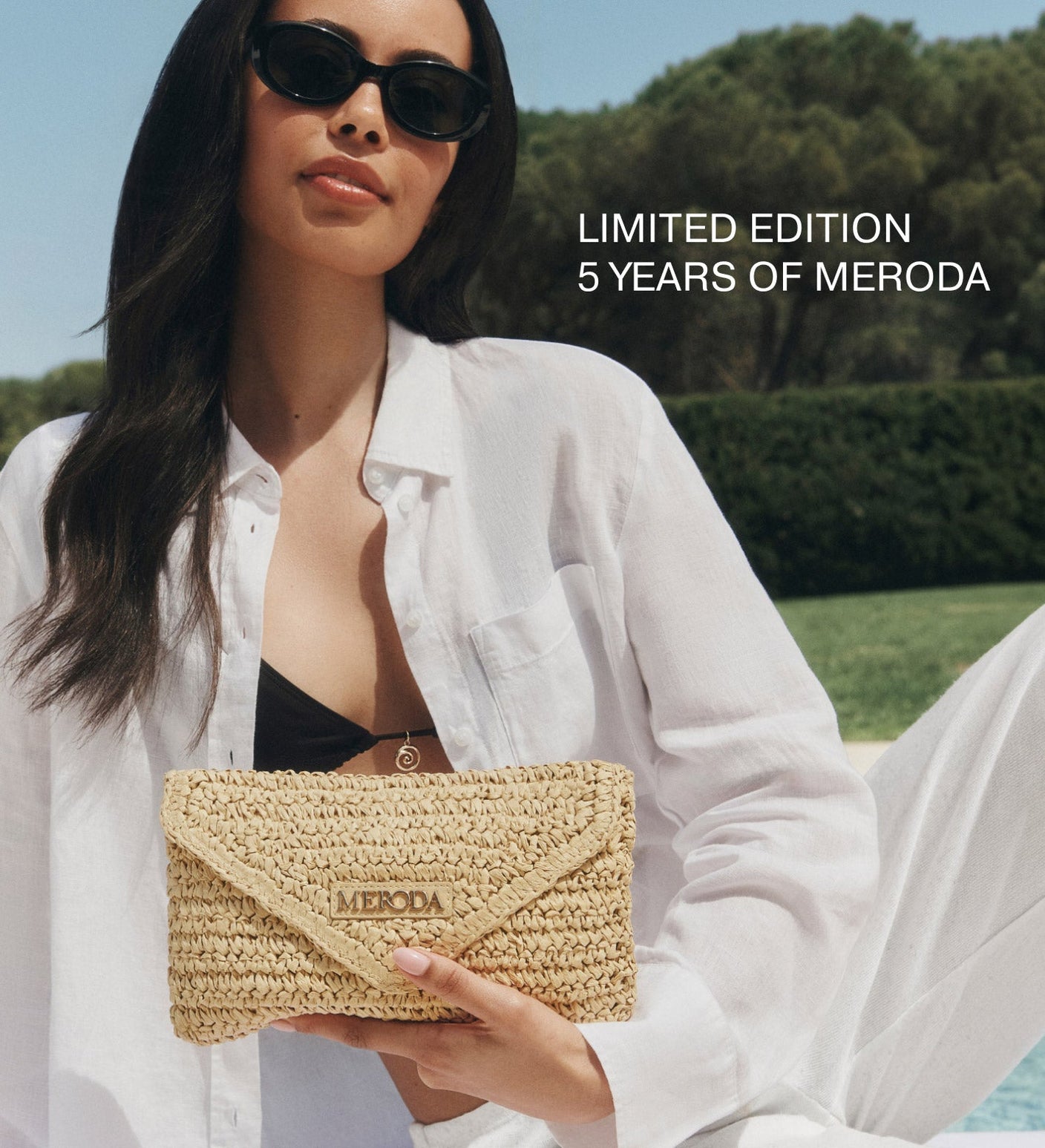 Summer Travel Clutch in woven paper straw held by a model wearing sunglasses in a sunny outdoor setting.